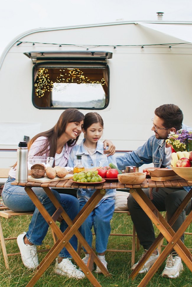 Familie isst am Tisch im Freien, mit einem Wohnwagen im Hintergrund.